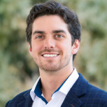 A smiling man with dark curly hair, wearing a white shirt and navy blazer, outdoors with greenery in the background.