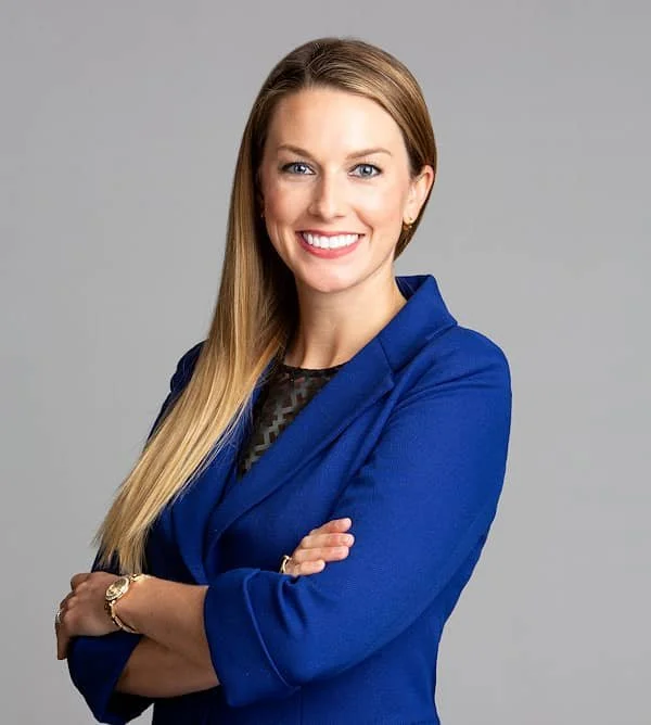 Professional woman with long blonde hair wearing a blue blazer, smiling with arms crossed, against a gray background.