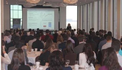 A large audience attending a conference or seminar in a spacious room with big windows, facing a speaker and a presentation screen displaying charts and data.