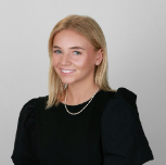 A young woman with blonde hair and a warm smile, wearing a black top and necklace, posing against a plain gray background.