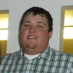 A young man with short dark hair and a goatee, smiling, wearing a plaid shirt.