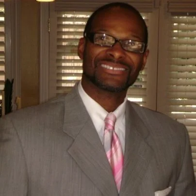 A man wearing glasses, a gray suit, white shirt, and pink striped tie, smiling indoors near window blinds.