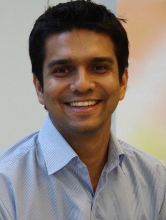 Smiling man with dark hair wearing a light blue collared shirt, posing indoors against a blurred background.