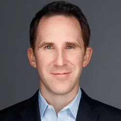 Professional portrait of a man with short brown hair, wearing a suit and a light blue shirt, smiling against a gray background.