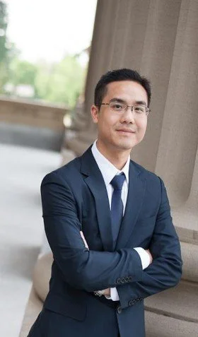 A young man in a dark blue suit and glasses standing outdoors, crossing his arms, near a stone structure with trees in the background.