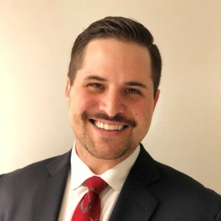 Portrait of a man with short dark hair, a mustache and beard, smiling, dressed in a suit with a white shirt and red patterned tie, standing in front of a plain light-colored background.