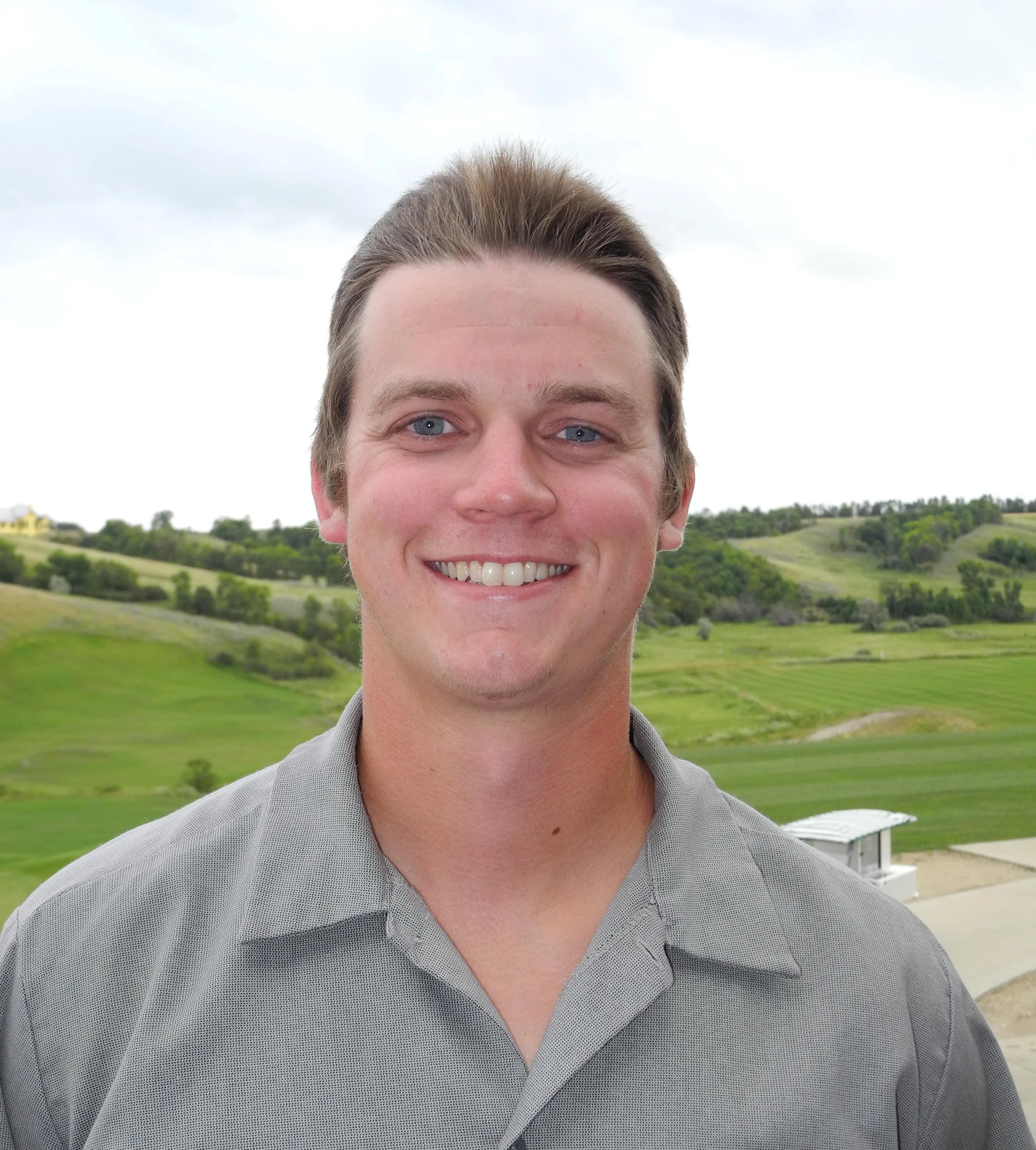 A young man with short brown hair and blue eyes smiling outdoors with a green hilly landscape and cloudy sky in the background.