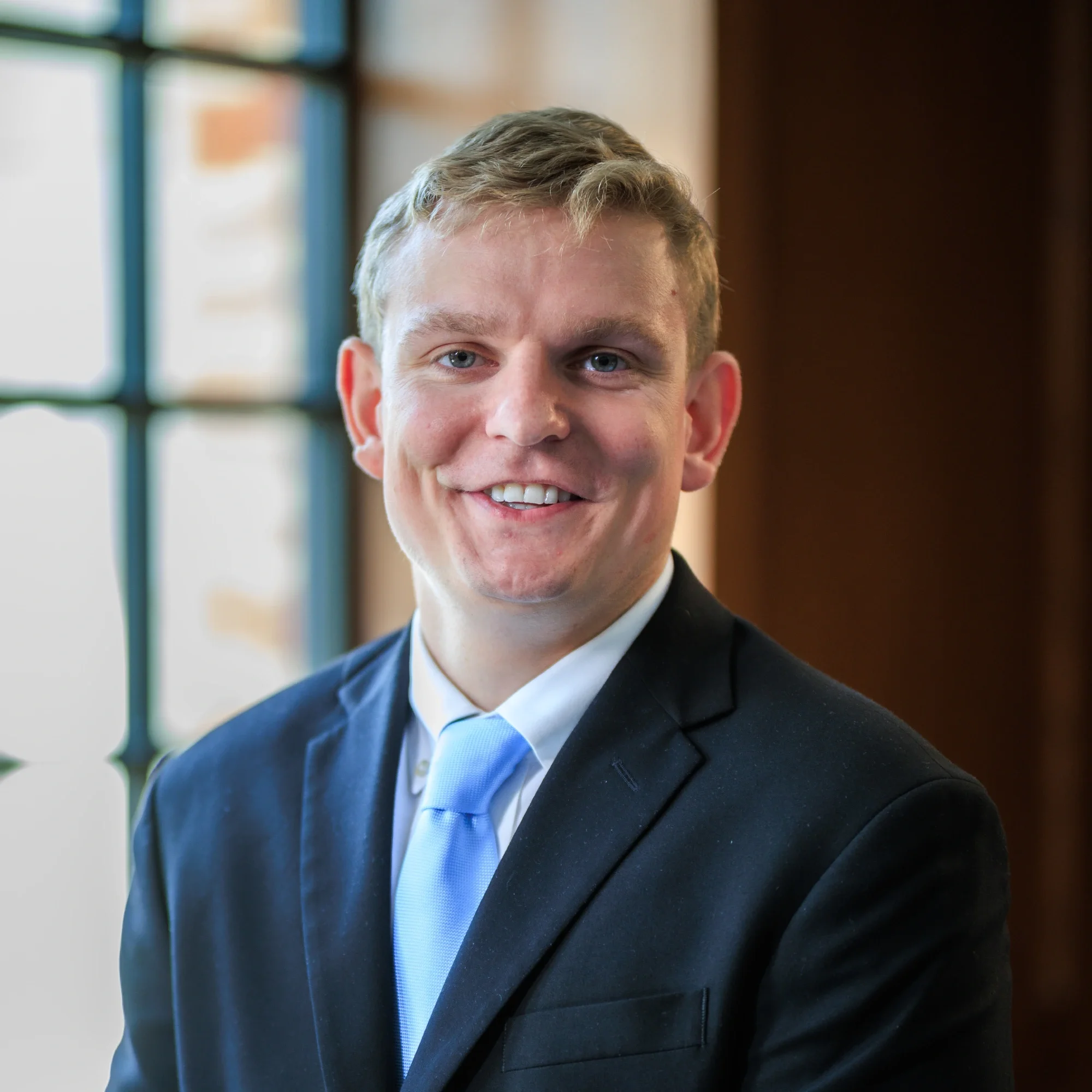 A young man in a business suit with a light blue tie, smiling, in an indoor setting with large windows in the background.