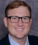 A headshot of a man with glasses and short, light brown hair, smiling against a dark background.