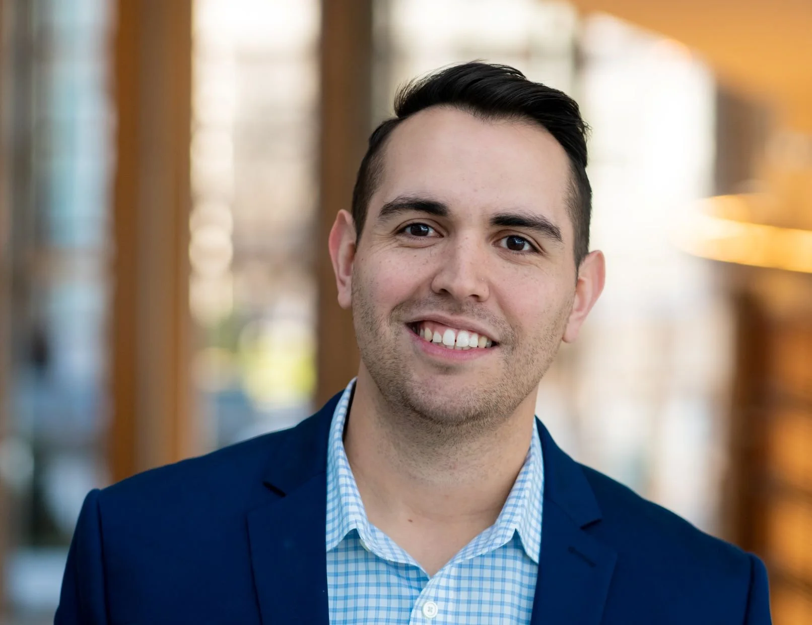 Portrait of a smiling man in a blue suit and checkered shirt, standing indoors with blurred background.
