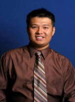 Portrait of a man with short black hair wearing a brown shirt and patterned tie, smiling against a blue background.