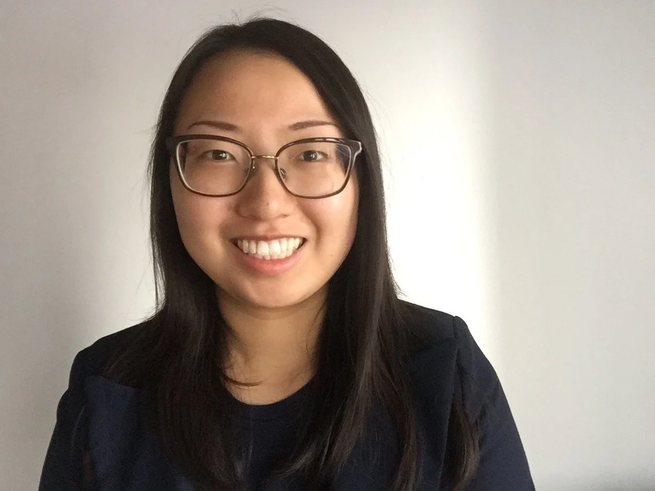A woman with long dark hair, wearing glasses, smiling against a plain light-colored wall.
