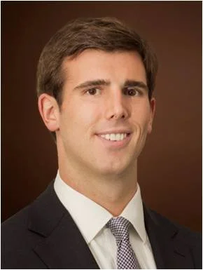 A professional headshot of a man with dark brown hair, wearing a black suit, white shirt, and a patterned tie, smiling against a brown background.