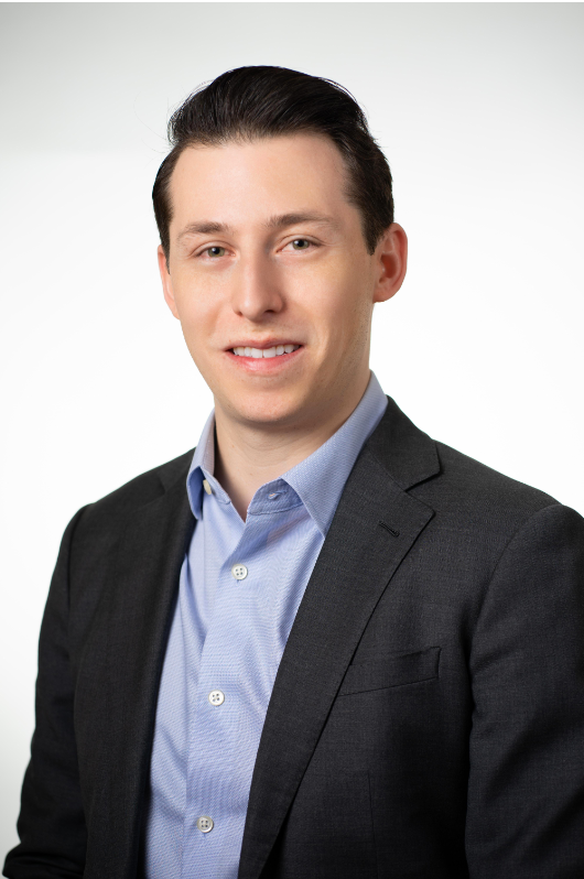 A young man with dark hair dressed in a dark suit and light blue button-up shirt, smiling at the camera against a plain white background.