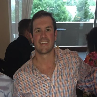 A smiling man wearing a plaid shirt at an indoor event with others in the background and large windows showing trees outside.