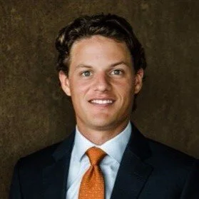 Portrait of a young man in a suit with an orange tie, smiling against a dark background.