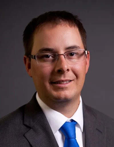 Professional headshot of a man wearing glasses, a navy blue suit, white shirt, and blue tie, against a gray background.