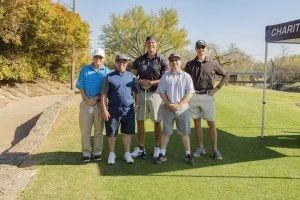 Six boys standing on a golf course near a flag that says 'Charity' in the background, with trees and a clear sky.