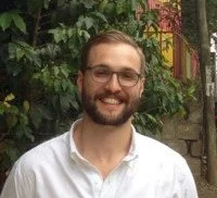 A smiling young man with glasses and a beard, wearing a white shirt, standing outdoors with green foliage and a wooden structure in the background.