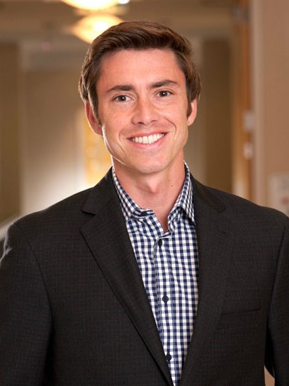 A young man with brown hair, wearing a black suit jacket and a checkered shirt, smiling and standing in a hallway.