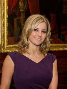 A woman with shoulder-length blonde hair, wearing a purple dress, and smiling, standing in front of a painting in an indoor setting.