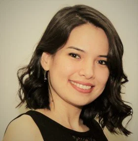 Portrait of a young woman with dark wavy hair smiling, wearing a black top and earrings, against a neutral background.