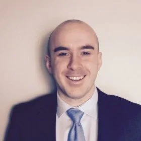A young man with a bald head, wearing a dark suit, white shirt, and light blue tie, smiling at the camera against a plain light-colored wall.