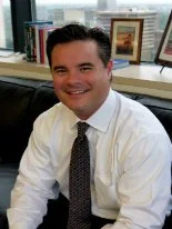 A smiling man sitting at a desk in an office, wearing a white shirt and tie, with books and framed pictures in the background.