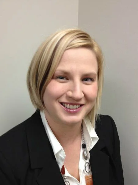 Portrait of a smiling woman with blonde hair wearing a black blazer and white shirt, standing against a plain wall.