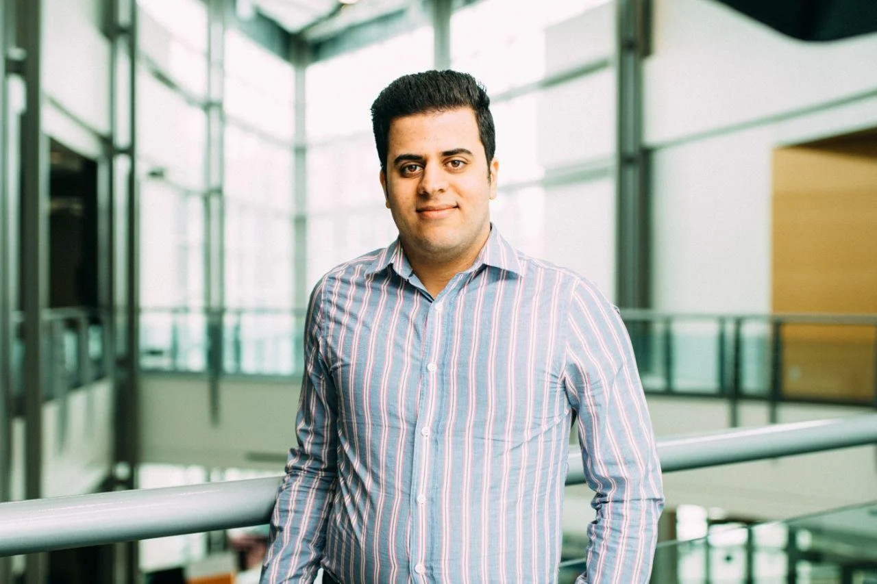 A young man in a striped button-up shirt standing in a modern building with large windows and metal railings, looking at the camera.