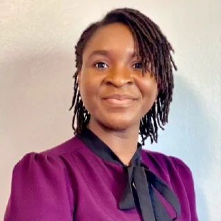 A woman with short dreadlocks wearing a purple top and black necktie, smiling at the camera.
