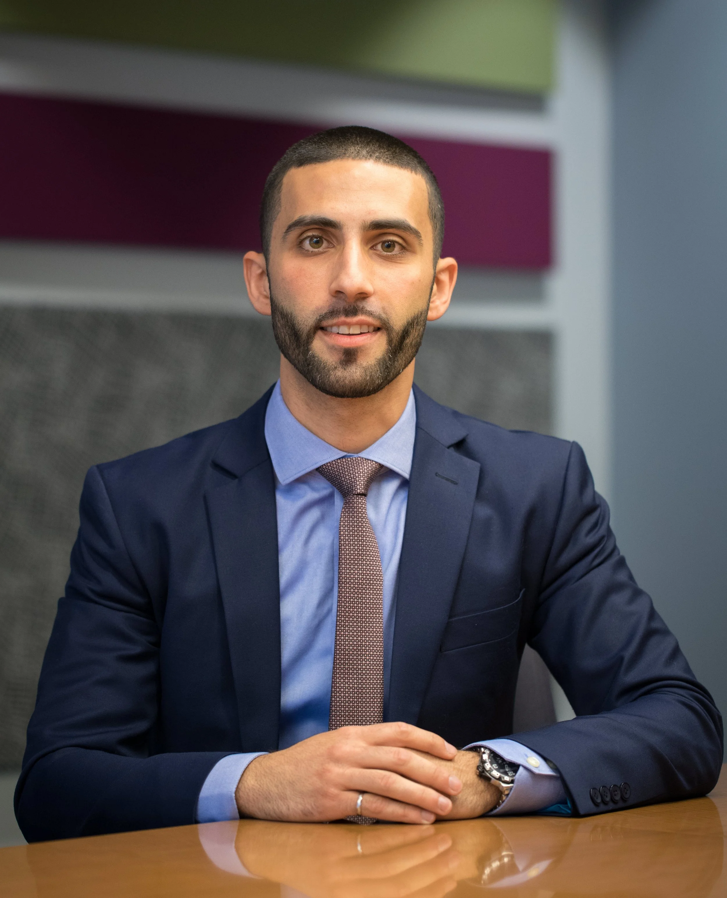 A man with short dark hair and a beard, wearing a navy blue suit, light blue dress shirt, and a pink tie, sitting at a wooden table in an office or conference room with colorful abstract wall art in the background.