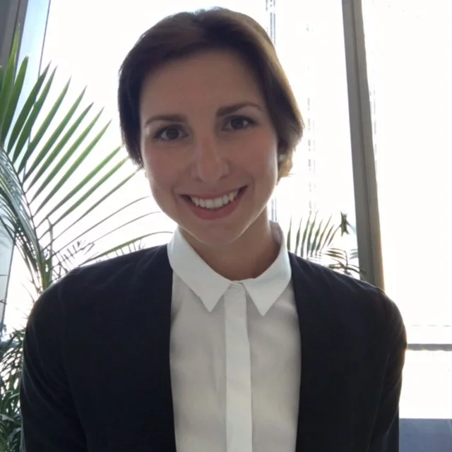 A woman with short brown hair smiling, wearing a white collared shirt and dark blazer, standing indoors near large windows and green plants.
