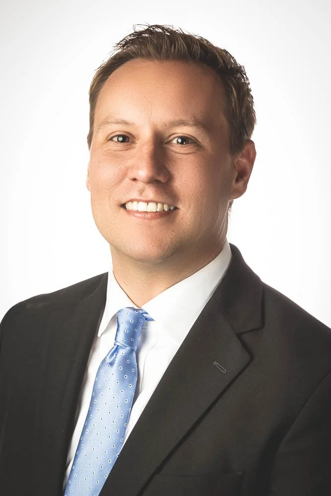 Professional headshot of a man in a black suit, white shirt, and light blue tie, smiling against a white background.
