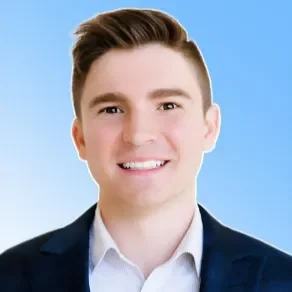 Portrait of a young man with short brown hair, wearing a navy blazer and white shirt, smiling against a light blue background.
