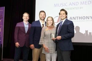 Four people standing on stage at an award event, with a projection screen behind them displaying 'Music and Media' and 'Presented by' and a name. One person is holding an award trophy.