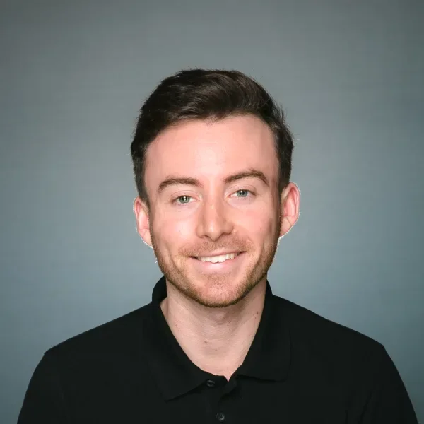 Headshot of a man with short dark hair, blue eyes, and light skin, wearing a black collared shirt, smiling against a gray background.