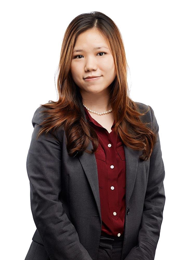 A young woman with long brown hair, wearing a dark gray blazer, a red blouse, and a pearl necklace, posing against a white background.