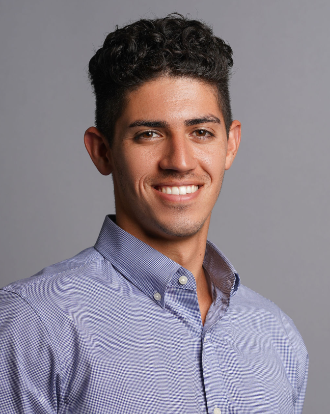 A young man with short, dark, curly hair smiling, wearing a light blue button-up shirt against a gray background.