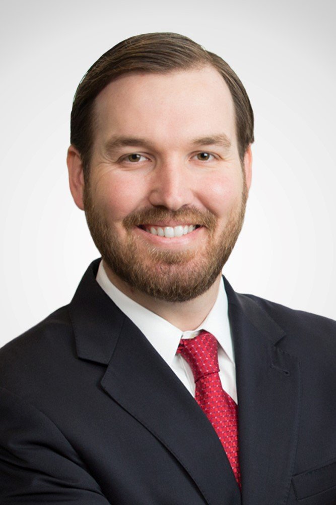 A professional headshot of a smiling man with brown hair and a beard, wearing a dark suit, white shirt, and red tie, against a plain white background.