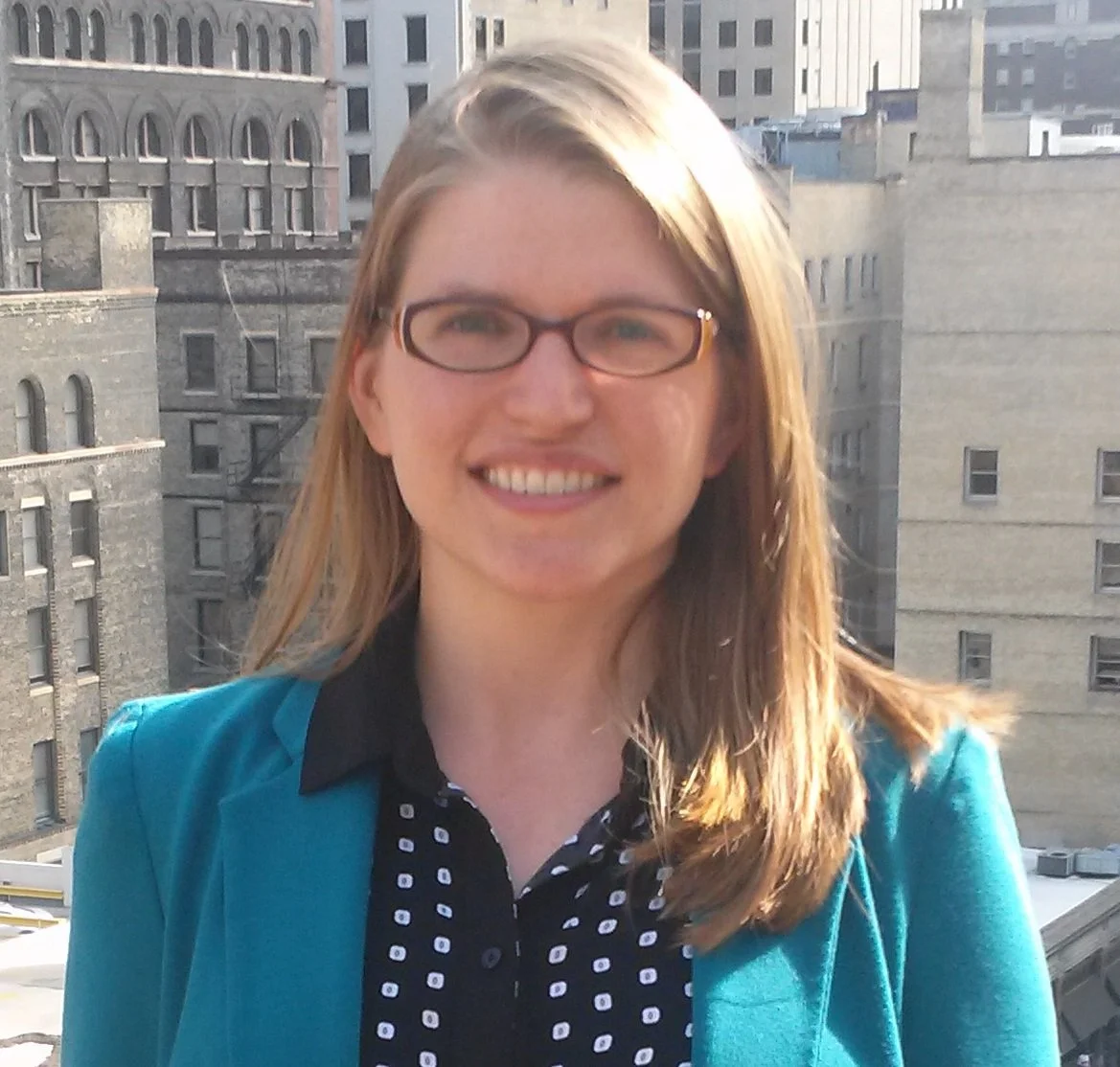 A woman with long blonde hair and brown glasses smiling outdoors in a city environment, wearing a teal blazer and a black-and-white polka dot shirt.