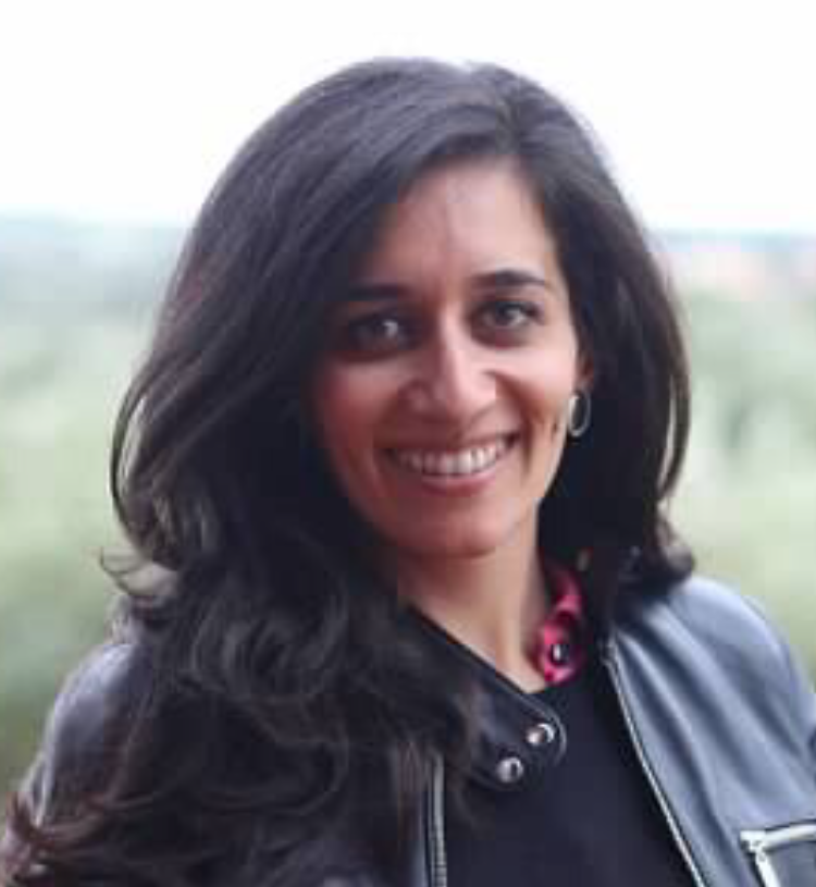 A woman with dark, shoulder-length hair smiling outdoors, wearing a leather jacket and hoop earrings.