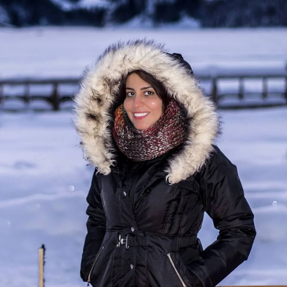 Woman smiling outdoors in winter, wearing a black parka with a fur-lined hood and a multicolored scarf, snow-covered landscape in the background.