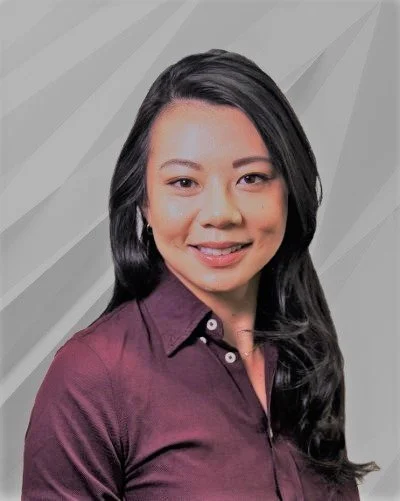 Portrait of a woman with long black hair, wearing a dark purple button-up shirt, smiling, against a gray background with textured diagonal lines.