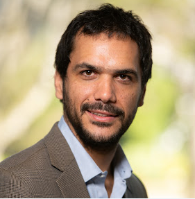Portrait of a man outdoors with a blurred green background, wearing a brown blazer and blue shirt.