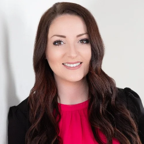 A young woman with long, wavy brown hair, wearing a black jacket over a bright pink top, smiling at the camera against a neutral background.
