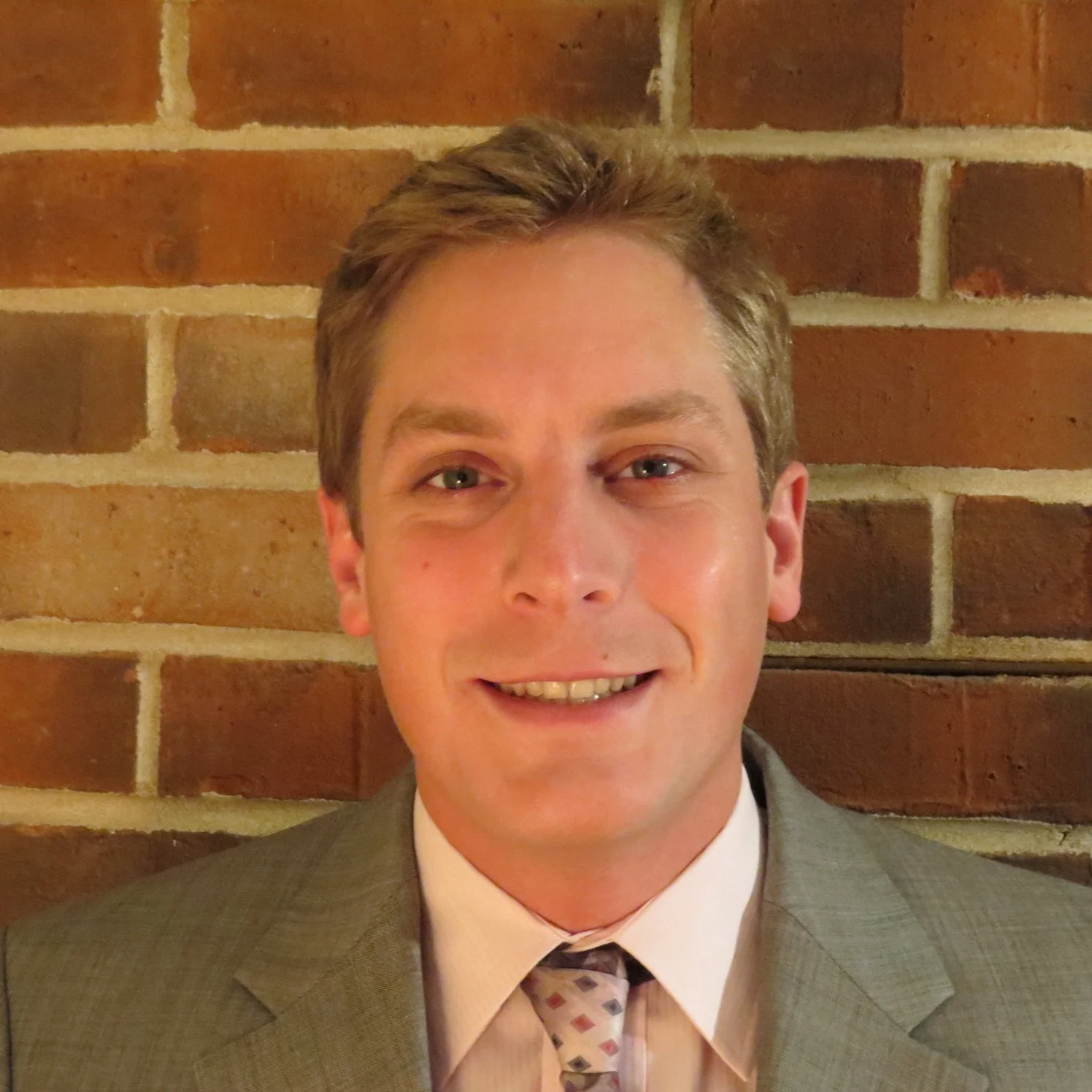 Portrait of a smiling man in a suit and tie in front of a brick wall.