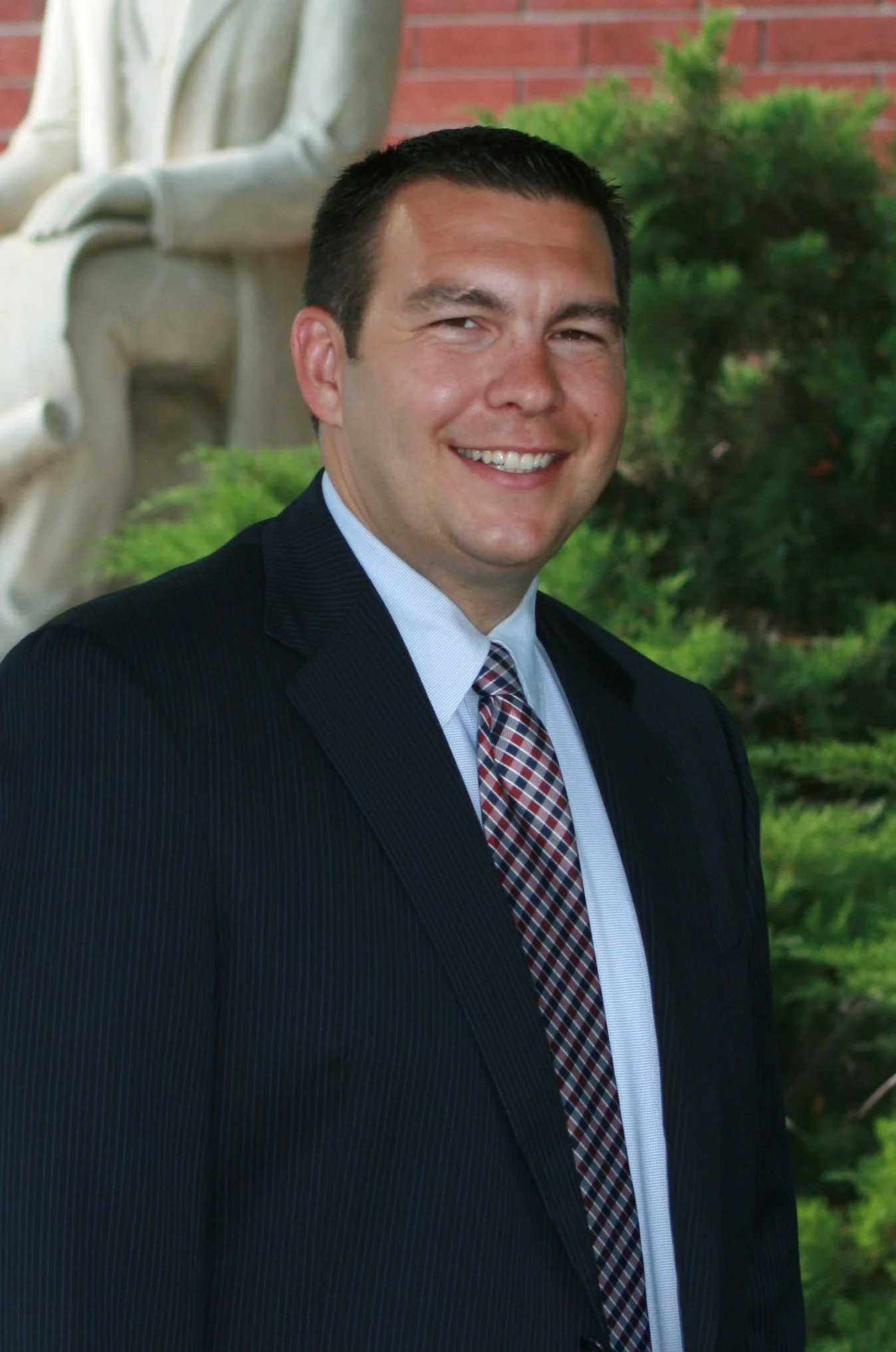 A man in a dark pinstripe suit with a light blue shirt and a plaid tie, smiling outdoors, with greenery and a sculpture in the background.