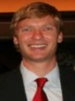A smiling man with light brown hair wearing a suit and red tie, standing against a dark background.