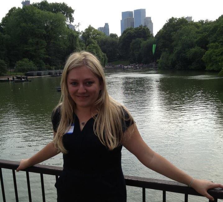 A young woman with long blonde hair standing by a river with trees on both sides and tall buildings in the background.